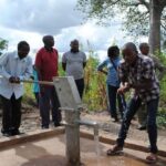 Yahaya Namangaya, Ndomoni village chairperson, pumps water for resident George Magomo, while members of village council discuss the borehole with PWRDF staff member Zaida Bastos (middle). Photo: André Forget