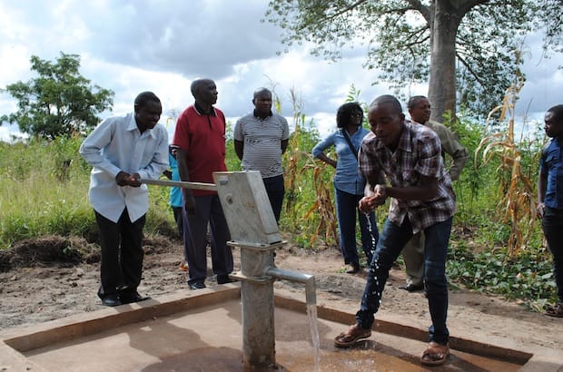 Yahaya Namangaya, Ndomoni village chairperson, pumps water for resident George Magomo, while members of village council discuss the borehole with PWRDF staff member Zaida Bastos (middle). Photo: André Forget
